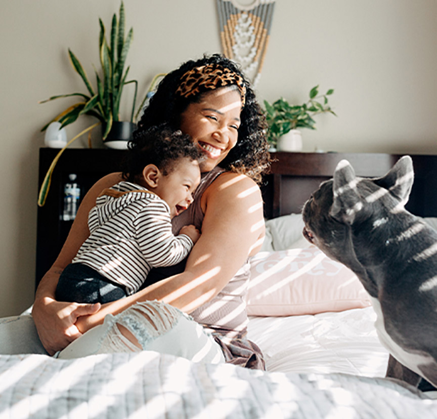 A woman and her child laughing in a sunny bedroom with their dog.
